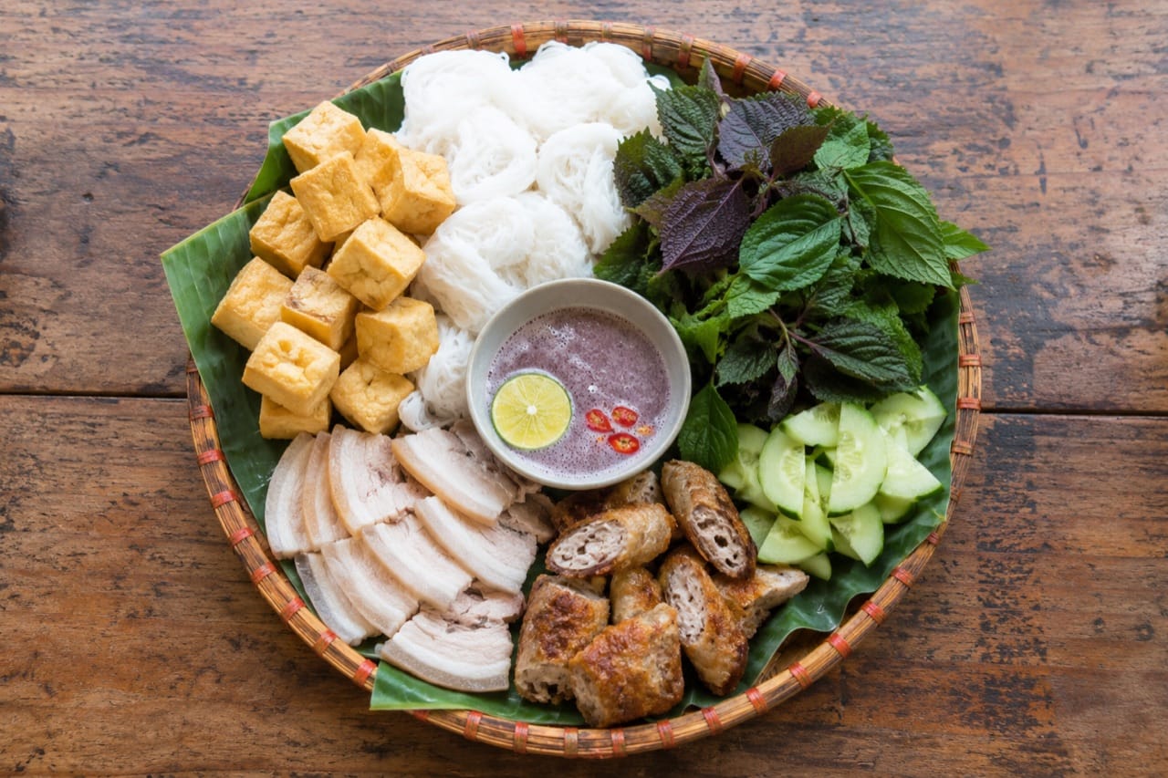 Mẹt tre lót lá chuối bày bún lá cắt khoanh, đậu chiên vàng, thịt luộc, chả chiên, rổ rau kinh giới tía tô và chén mắm tôm đánh bọt