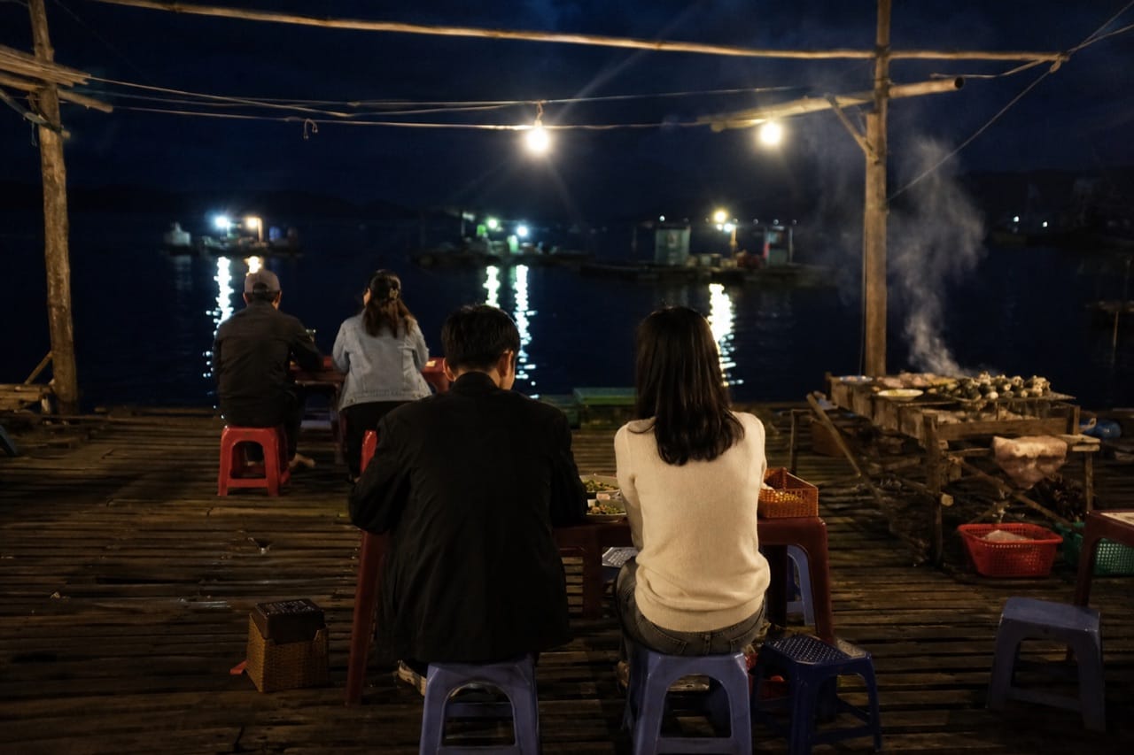 Quán bè trên mặt biển ban đêm, bóng đèn tròn treo trên đầu, bàn ghế nhựa kê trên sàn ván, xa xa là đèn thuyền